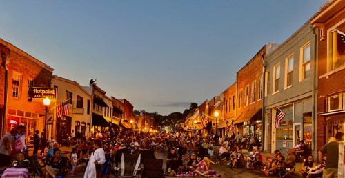 A bustling street scene at dusk, filled with people enjoying a lively outdoor gathering among shops and lights.