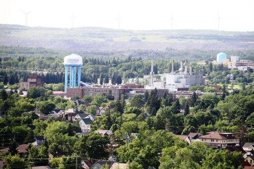 Aerial view of a town with a water tower, industrial buildings, and wind turbines in the background, surrounded by greenery.