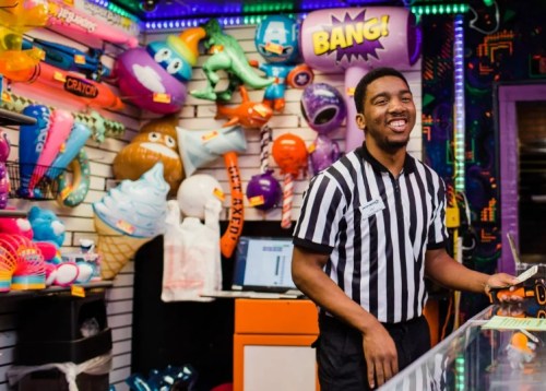 A smiling store employee in a striped shirt stands behind a counter filled with colorful toys and decorations.