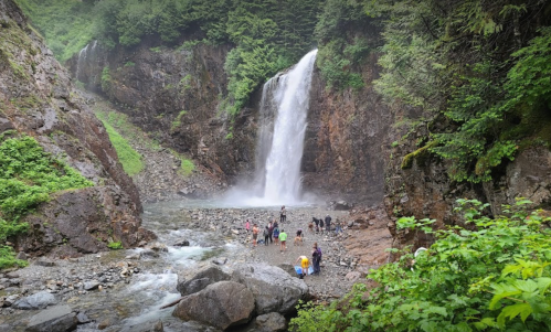 A waterfall cascades down rocky cliffs, surrounded by lush greenery and people exploring the area below.