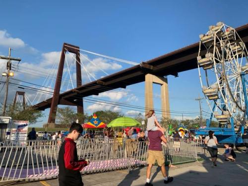 A lively fair scene with a ferris wheel and a bridge overhead, featuring people enjoying the festivities.