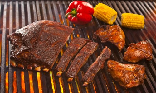 Grilled meats including ribs, chicken, and corn, with red bell pepper, on a barbecue grill.