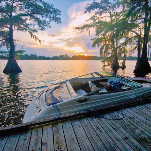 A boat docked on a wooden pier at sunset, surrounded by trees and calm water reflecting the colorful sky.