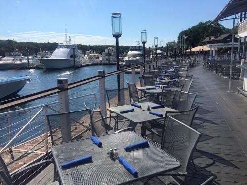 Outdoor dining area by a marina, featuring empty tables and boats docked in the water under a clear blue sky.