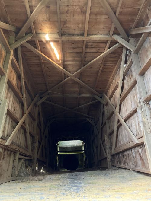 View of a wooden tunnel with a dark opening at the end, illuminated by a beam of light from above.