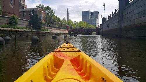 A yellow kayak on calm water, with buildings and a bridge in the background under a partly cloudy sky.