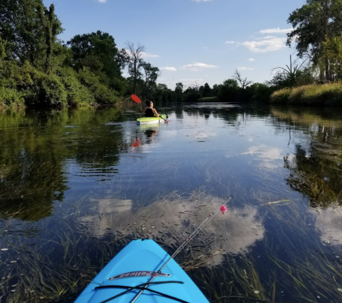A person kayaking on a calm river surrounded by trees and reflecting clouds, with a fishing rod visible in the foreground.