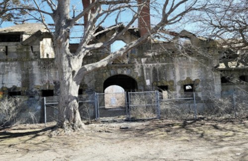 Abandoned building with crumbling walls, a large archway, and a bare tree in front, surrounded by a chain-link fence.