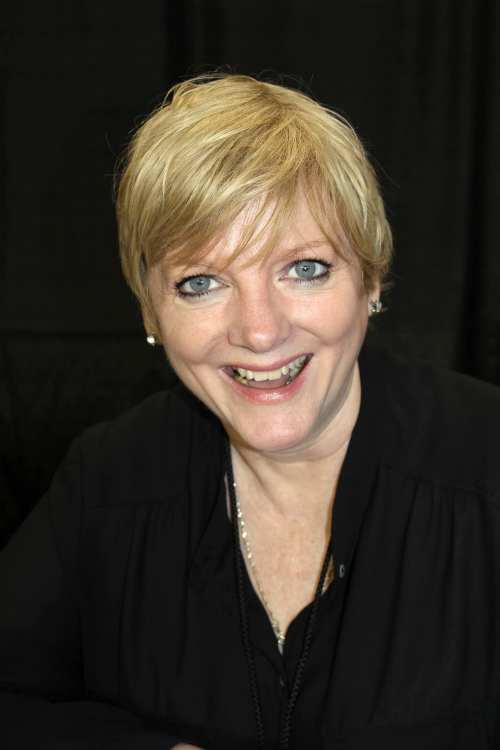 A smiling woman with short blonde hair, wearing a black top, against a dark background.