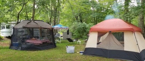 Two camping tents set up in a wooded area, with a small fire and outdoor furniture nearby.