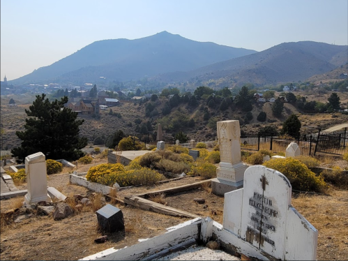 A cemetery with gravestones in the foreground, set against a mountainous landscape and a small town in the distance.