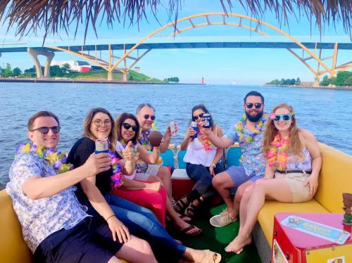 A group of seven friends on a boat, wearing leis and enjoying drinks, with a bridge and water in the background.