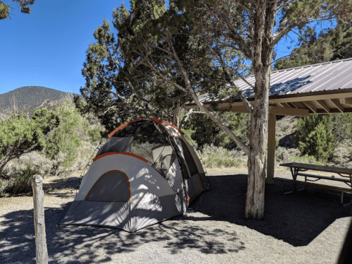 A tent set up under a tree near a picnic shelter in a scenic outdoor area with mountains in the background.