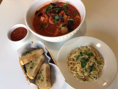 A bowl of seafood stew with herbs, a side of spaghetti, and toasted bread with parsley on a white table.