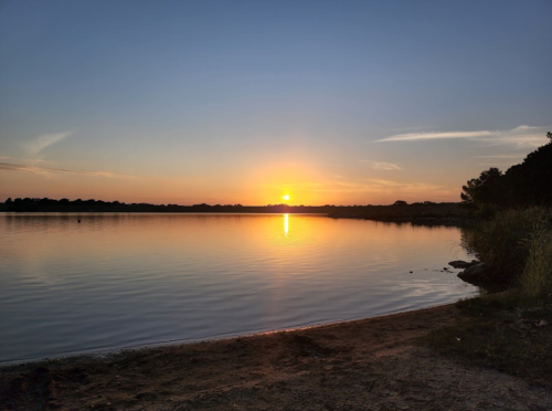 A serene sunset over a calm lake, with reflections on the water and silhouettes of trees along the shore.