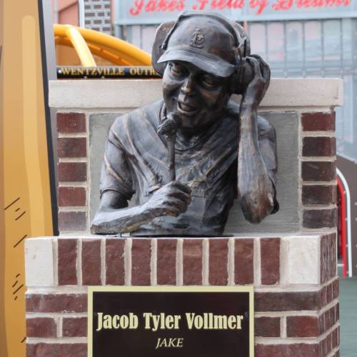A bronze statue of a smiling man wearing a cap and headphones, mounted on a brick pedestal with a plaque.