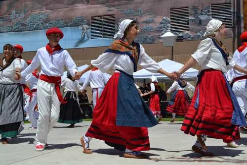 A group of dancers in traditional attire perform a lively dance, holding hands in a festive outdoor setting.