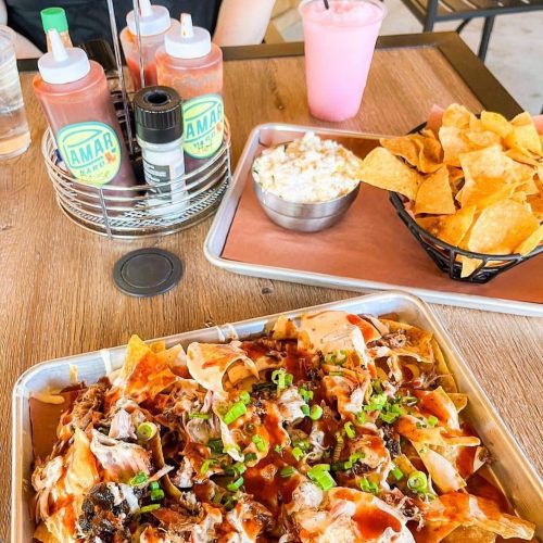 A tray of loaded nachos topped with green onions, alongside chips, a drink, and a bowl of coleslaw on a wooden table.
