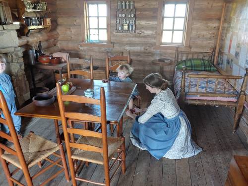 A woman in a blue dress tidies a wooden table in a rustic cabin, while two children play nearby.