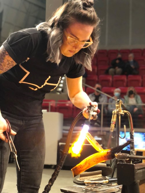 A person with glasses and tattoos uses a torch to work on a piece of art, with an audience in the background.