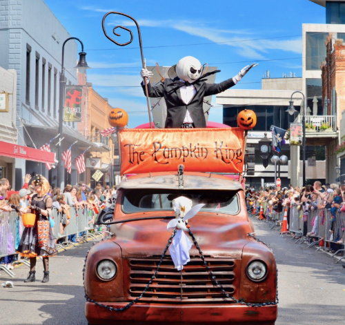 A festive parade scene featuring a character in a tuxedo on a vintage truck, surrounded by Halloween decorations and spectators.
