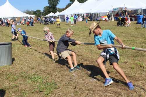 Children participate in a tug-of-war game at an outdoor event, with tents and spectators in the background.