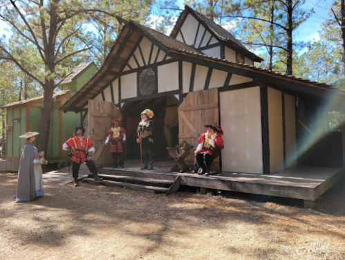 A group of people in historical costumes stands outside a rustic wooden building in a forested area.