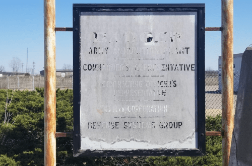 Weathered sign for the Army Ammunition Plant, listing representatives and contractors, with faded text.