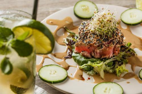 A plate of sesame-crusted tuna on greens, garnished with sprouts, cucumber slices, and a drizzle of sauce, with a drink nearby.