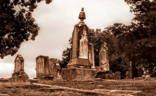 A sepia-toned image of old gravestones in a cemetery, surrounded by trees under a cloudy sky.