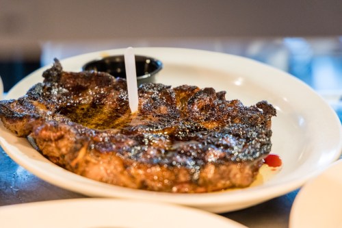 A grilled steak on a white plate, garnished with a small sauce cup and a toothpick.