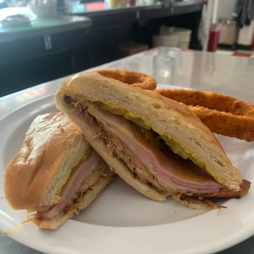 A close-up of a sandwich with layers of meat and pickles, served on a plate with crispy fries in the background.