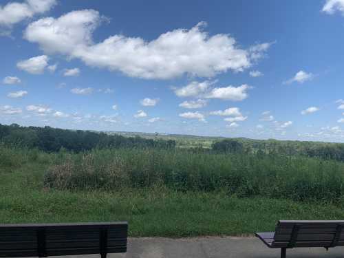 A scenic view of green hills and blue skies with fluffy clouds, framed by two empty benches in the foreground.