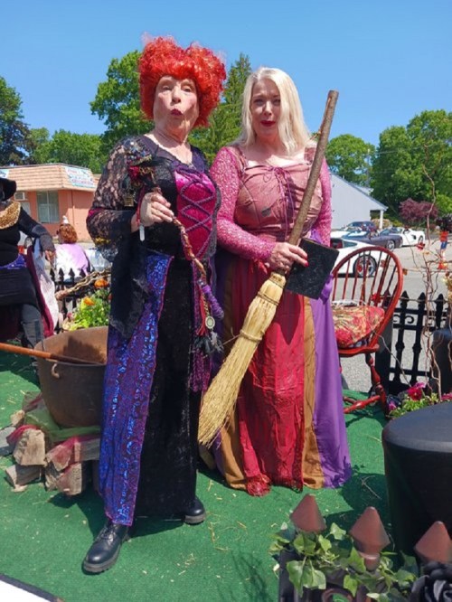 Two women in colorful, elaborate costumes stand together, one holding a broom, against a sunny outdoor backdrop.