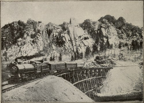 A vintage steam locomotive crosses a wooden bridge surrounded by rocky cliffs and trees.