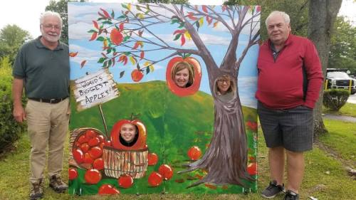 Four people pose behind a colorful apple-themed cutout, with a sign reading "World's Biggest Apples."