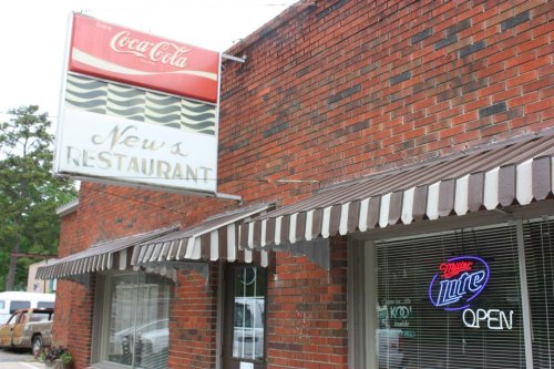 A brick building with a Coca-Cola sign and a neon "Open" sign in the window of New's Restaurant.