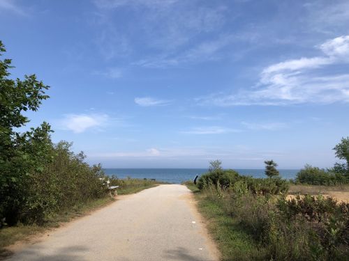 A sandy path leads to the ocean, surrounded by greenery and a clear blue sky with scattered clouds.