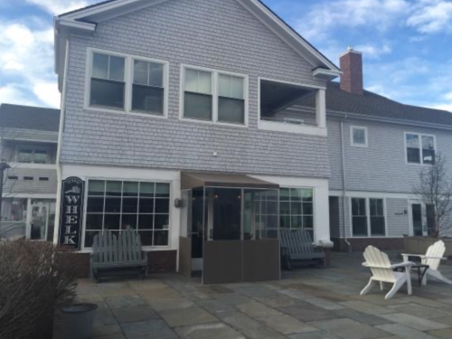 A gray shingle-style building with large windows and a patio featuring Adirondack chairs and a sign that reads "Whelk."