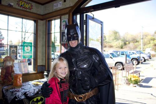 A child in a red outfit poses with a man in a Batman costume inside a cafe, smiling together.