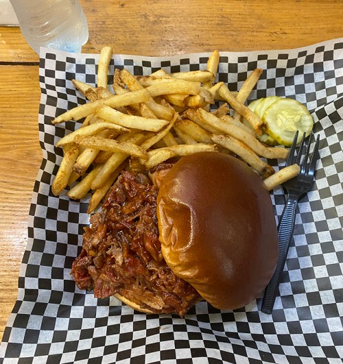 A pulled pork sandwich on a bun with fries and a pickle, served on a checkered paper plate.