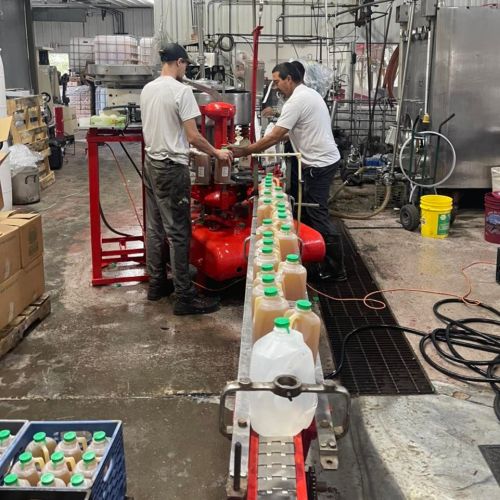 Two workers fill containers on a production line in a warehouse, with equipment and supplies in the background.