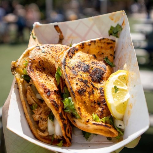 Two grilled tacos filled with meat and garnished with cilantro, served with a lemon wedge in a takeout container.