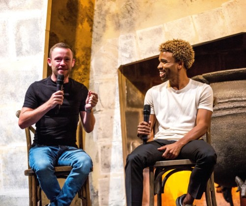 Two men sit on wooden chairs, speaking into microphones in a cozy, warmly lit setting.