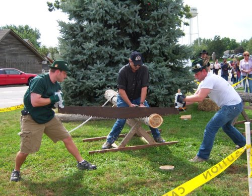 Two men saw a log together using a large hand saw, while a third man assists, with a tree and water tower in the background.
