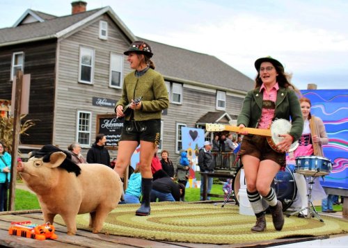 Two women perform on a stage with a pig statue, one playing a guitar and the other a banjo, in a lively outdoor setting.