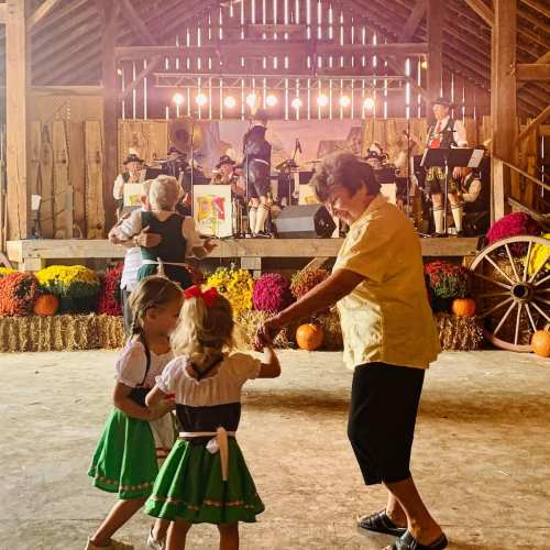 Two young girls in traditional dresses dance with an older woman in a barn, while a band performs in the background.