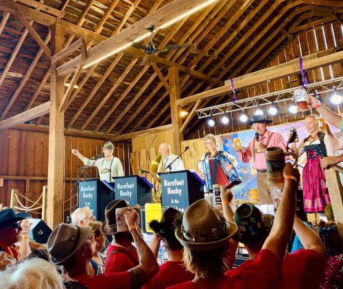 A lively barn event with performers on stage, audience cheering, and festive decorations in a rustic setting.