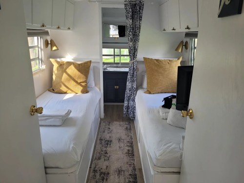 Cozy interior of a camper with two twin beds, light curtains, and a small kitchenette in the background.