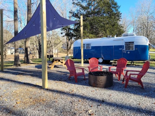 A cozy outdoor space with red chairs around a fire pit, a blue vintage trailer, and a shaded area under a purple canopy.